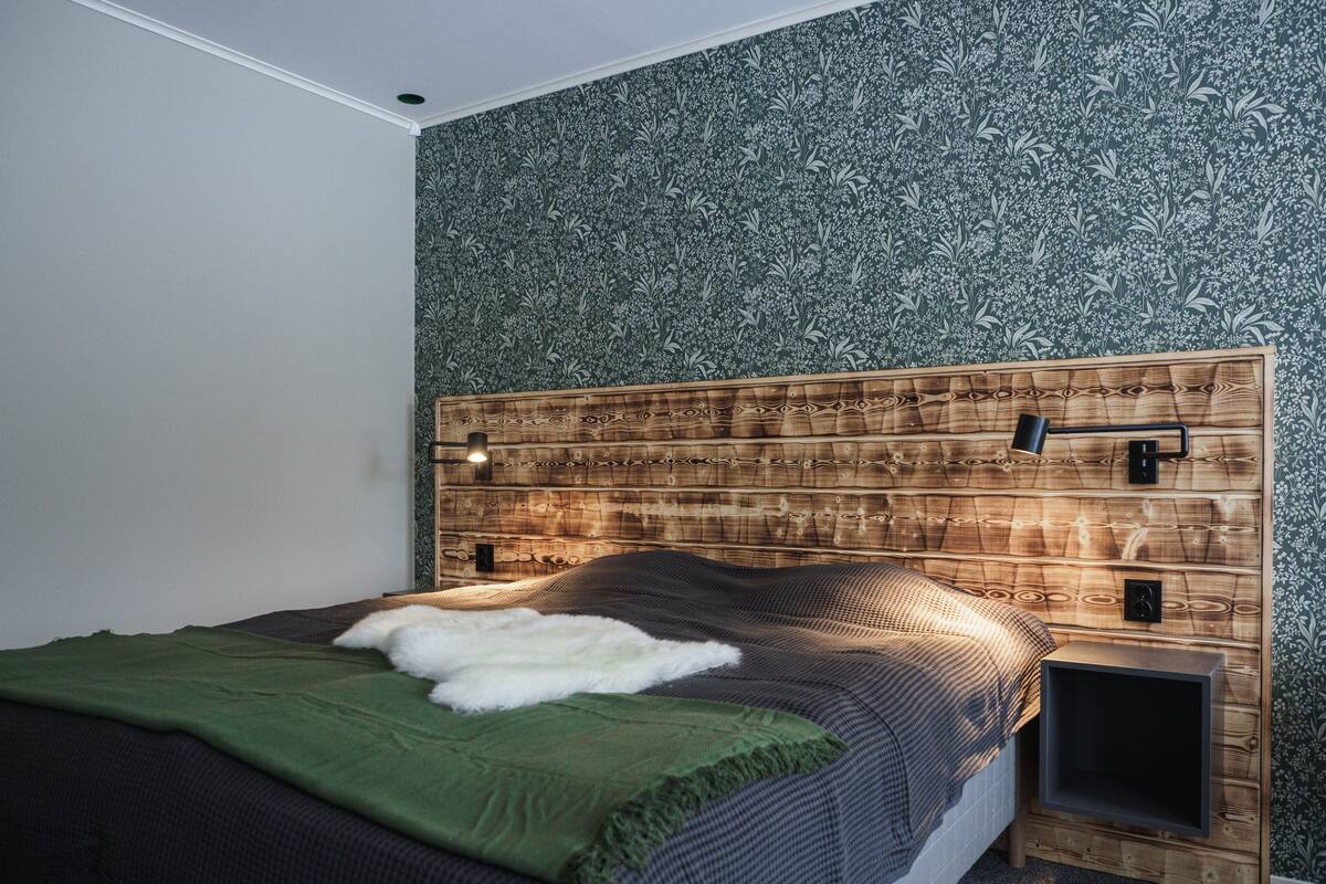Quarto com cabeceira de madeira, cama com cobertor verde e uma pequena mesa de cabeceira.