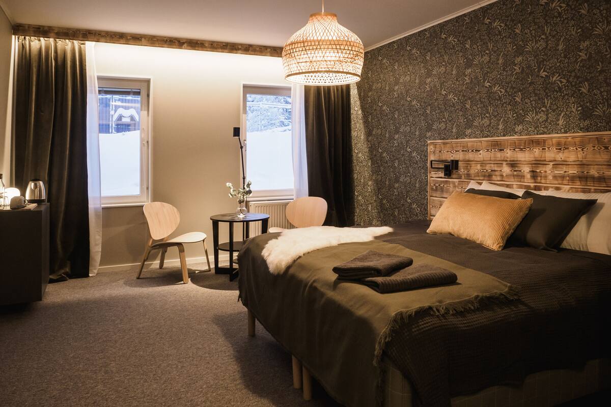 Quarto de hotel com cama, escrivaninha, cadeira e janela com cortinas.