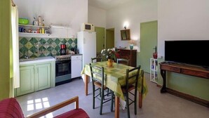 A compact kitchen with a stove, microwave, and refrigerator, a dining table with chairs, and a television on a wooden stand. - Château Teillan - Cadran Solaire (Aimargues)