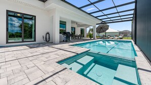 Een modern buitenzwembad met glazen omheining, een barbecue en een terras met lounge fauteuils.