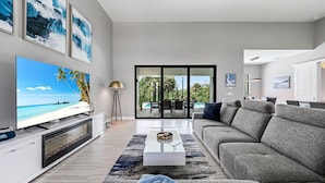 Een moderne woonkamer met een flatscreen televisie, een grijze bank, een bijzettafeltje en een groot raam met uitzicht op een zwembad en tuinmeubilair.