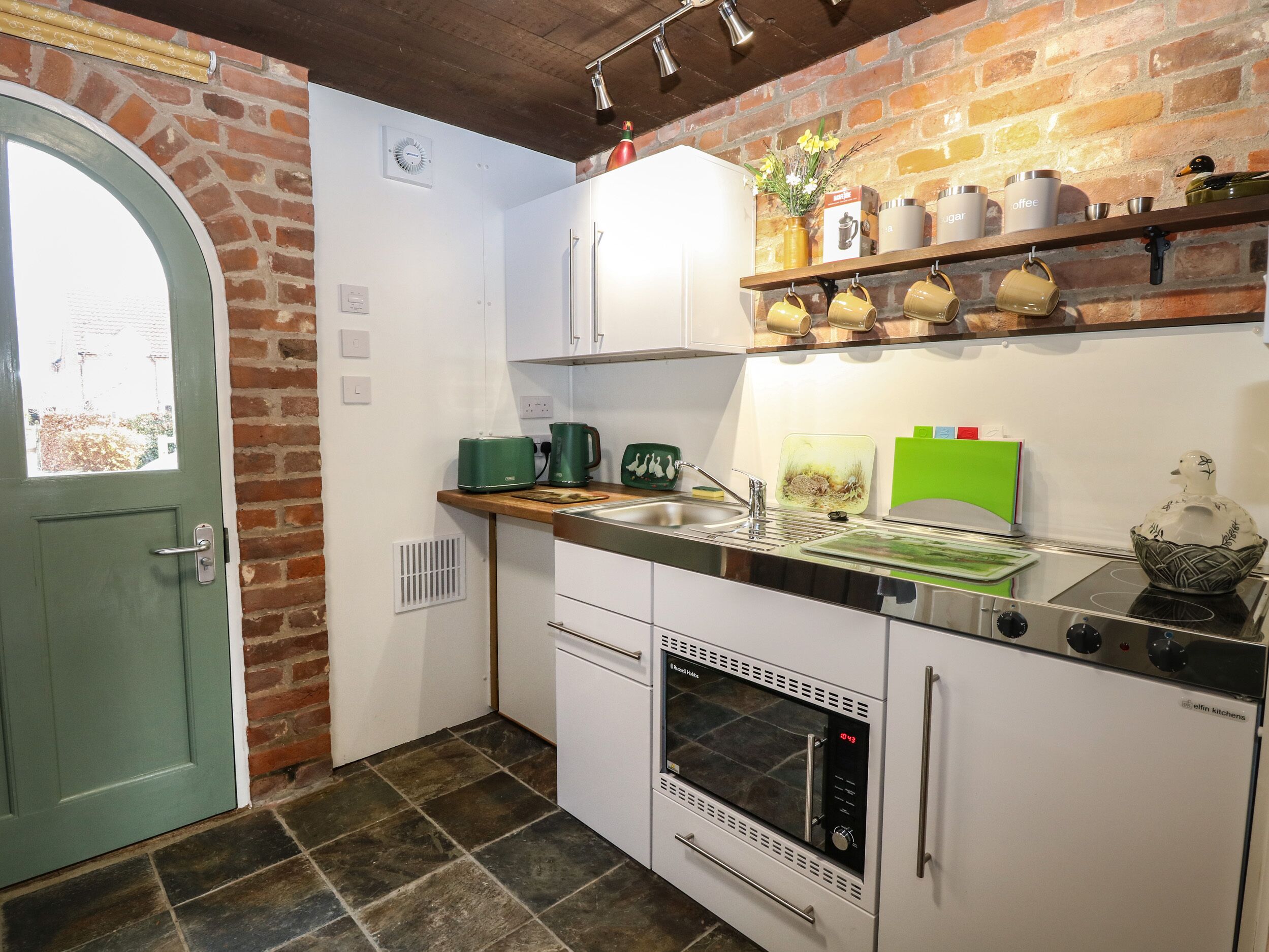 Une cuisine compacte avec un mur en briques, une porte verte, des placards blancs, un four encastré et une étagère contenant des ustensiles de cuisine.