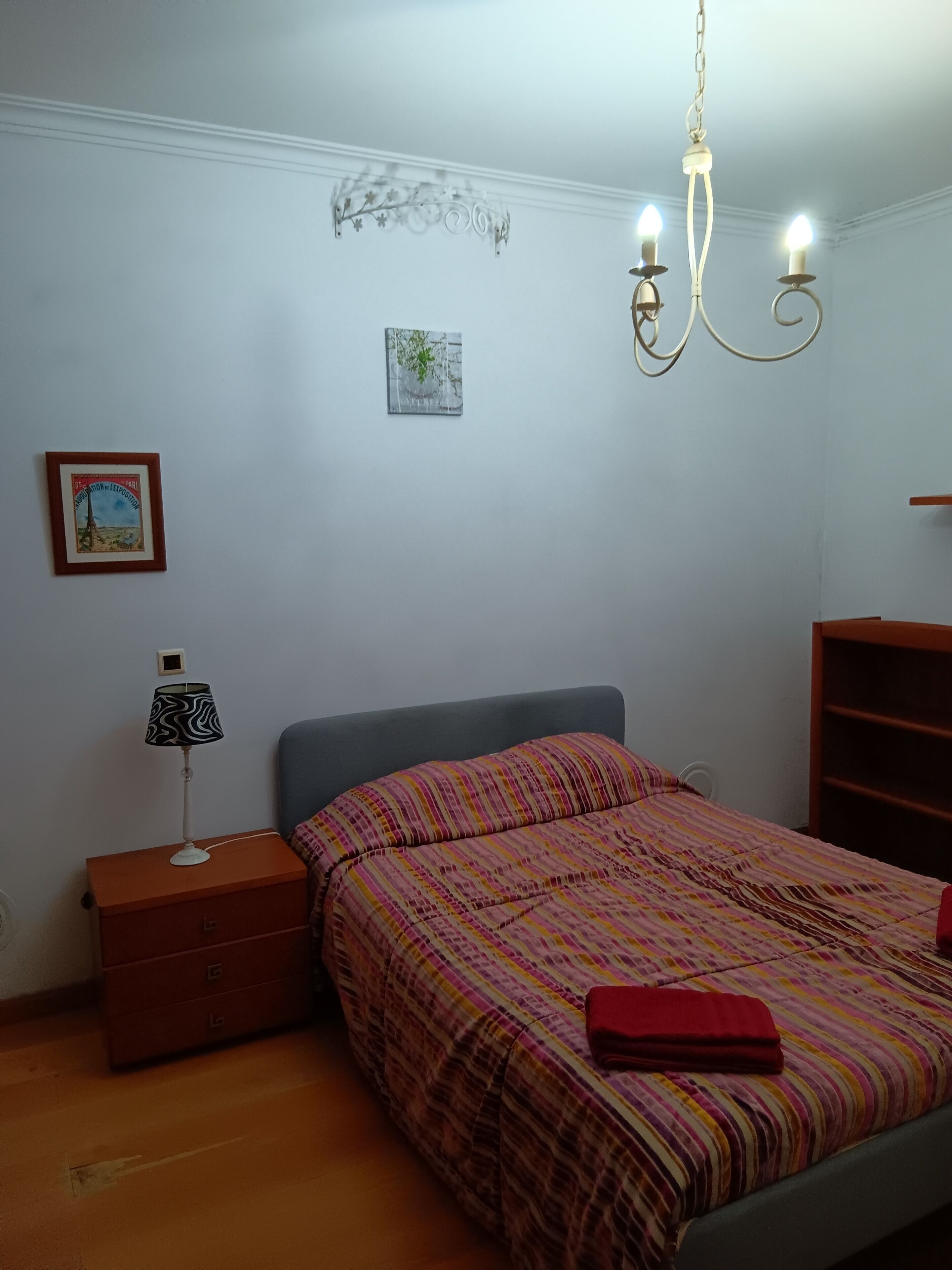 A bedroom with a bed, a nightstand, a lamp, a chandelier, and a wall with two pictures.