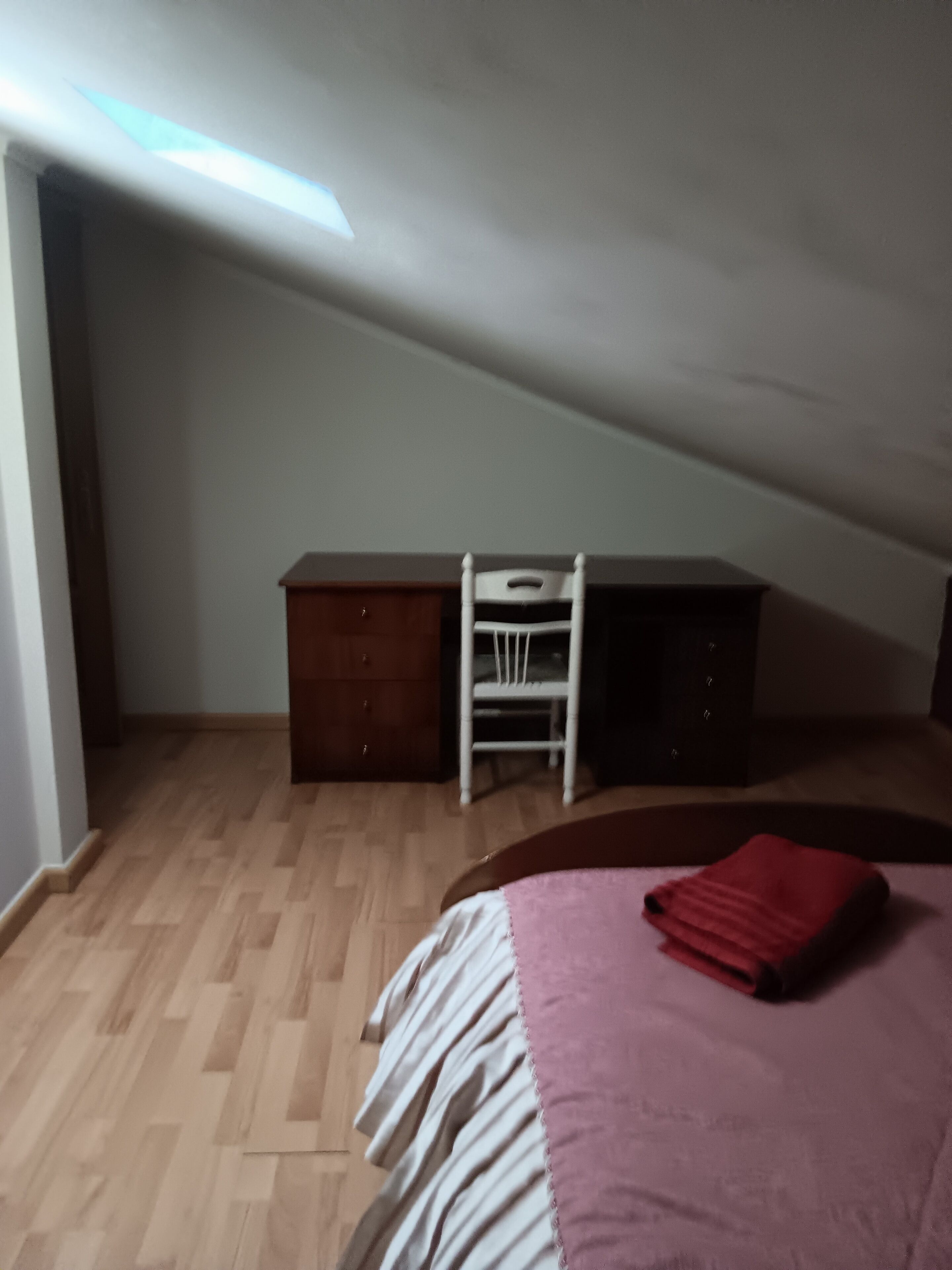 Een kamer met een schuine kap, een bed met een rode deken, een houten dressoir en een witte stoel.