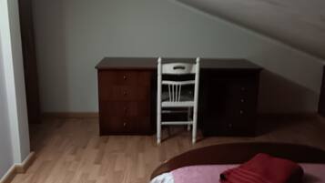 Een kamer met een schuine kap, een bed met een rode deken, een houten dressoir en een witte stoel.