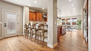 A spacious kitchen with wooden flooring, a central island with bar stools, wooden cabinets, and a dining area with a table and chairs. - Large Modern 5 Bedroom Custom Home (Meridian)