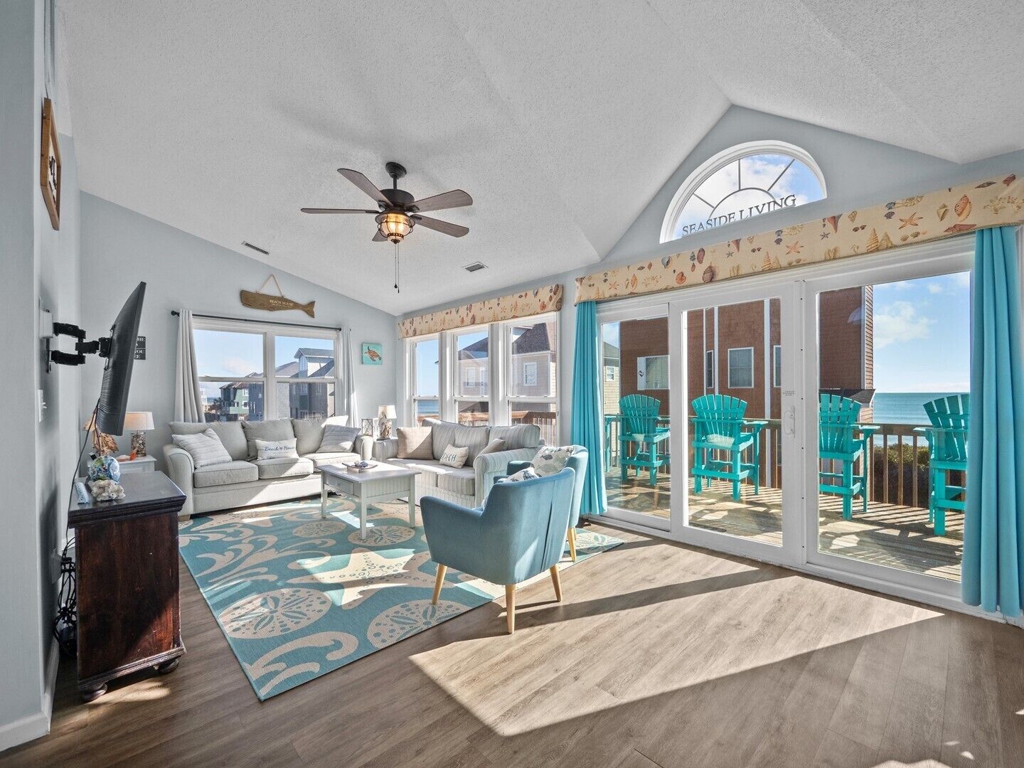 A spacious living room with a large sliding glass door leading to an outdoor deck, a ceiling fan, and a view of the sea.