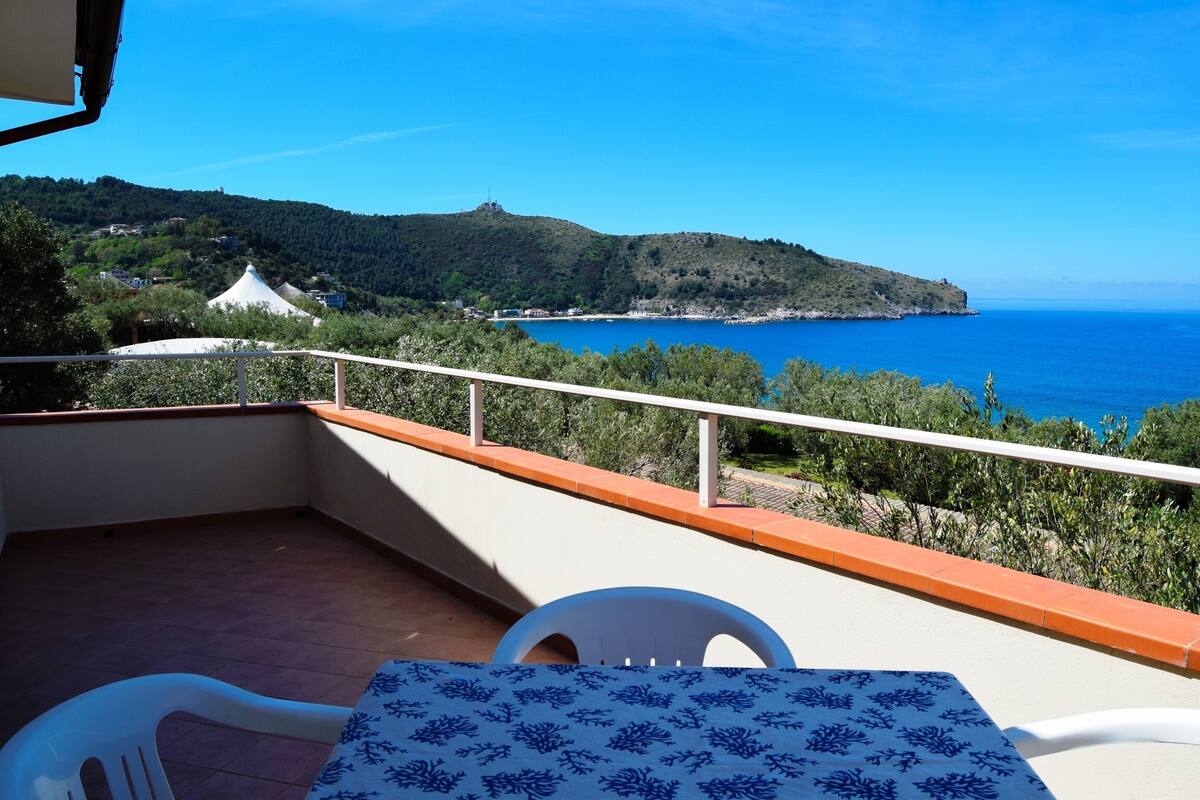 A balcony with a table and chairs, overlooking a coastal landscape with a hill and the sea.