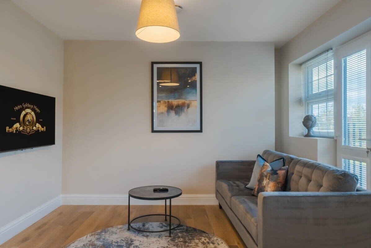 A living room with a sofa, a small round table, a hanging light fixture, a framed picture on the wall, and a window with blinds.