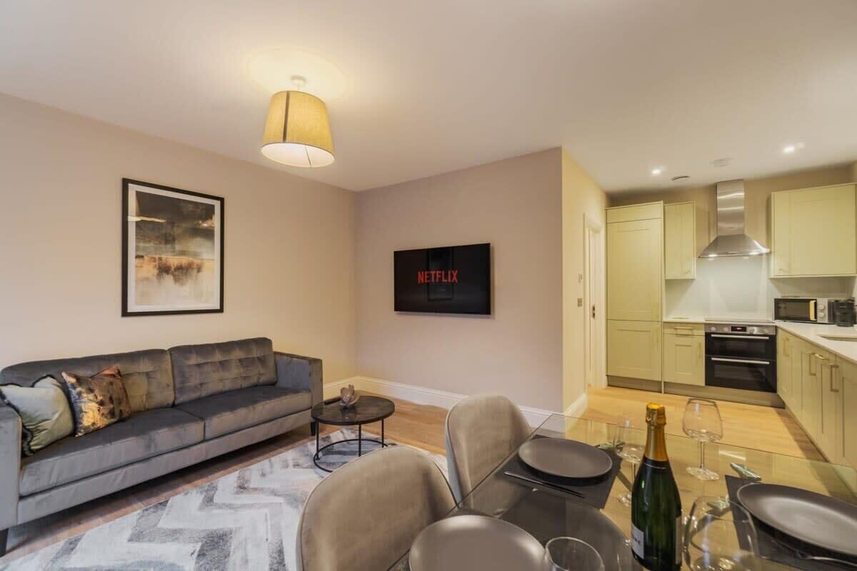 A modern living room with a sofa, a coffee table, and a dining area with chairs and a glass table. A TV and a framed picture are on the wall.