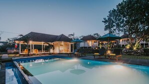 A swimming pool with lounge chairs and umbrellas, surrounded by houses with outdoor lighting. - Paluh Beach Huts (Lembongan Island)