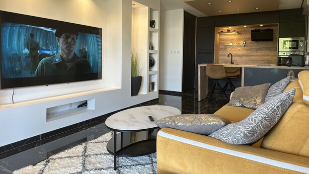 A modern living room with a flat-screen TV, a sofa, a coffee table, and a kitchen area in the background.