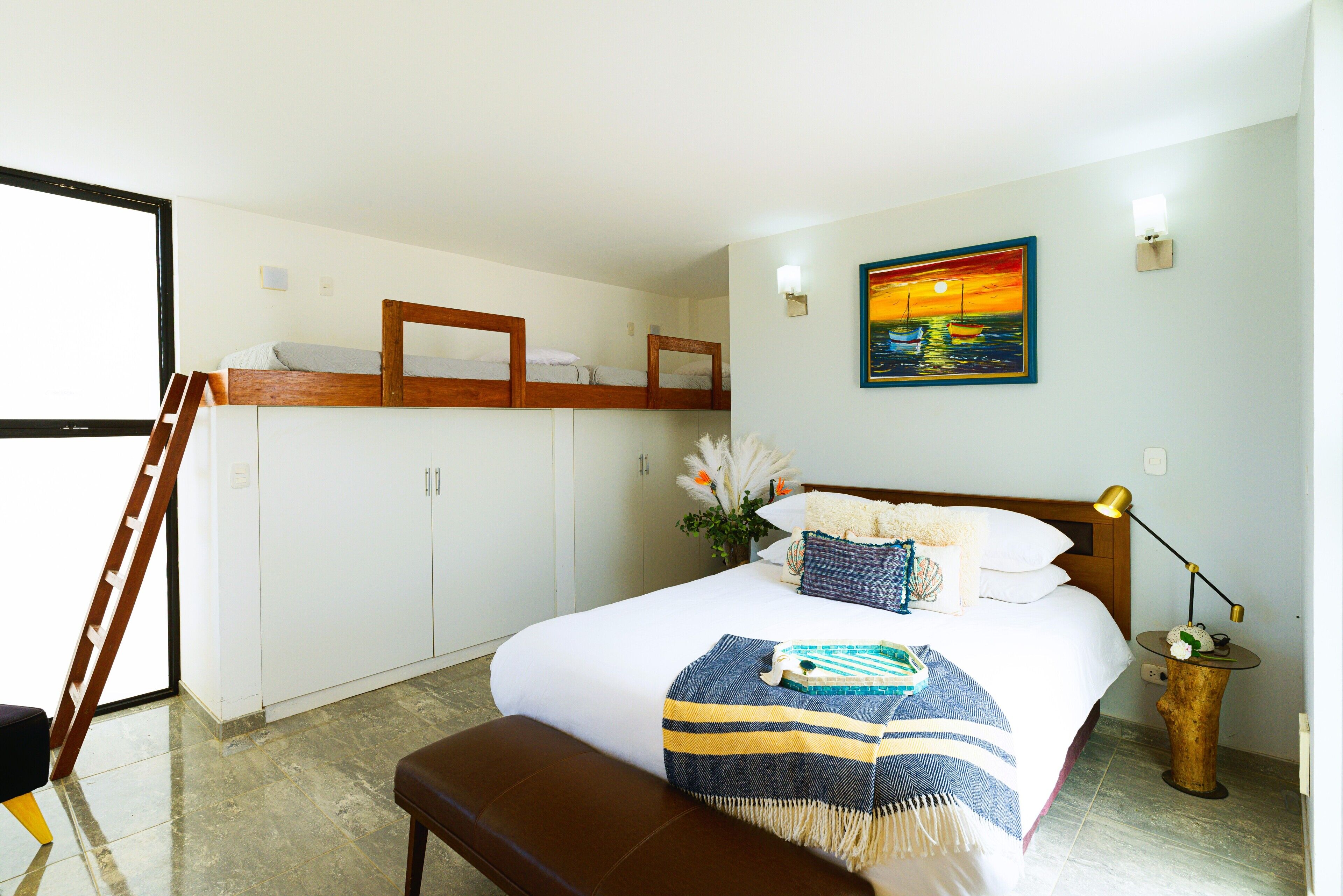 A bedroom with a large bed, a wooden headboard, a bench, a nightstand with a lamp, a painting, and a window with a view.