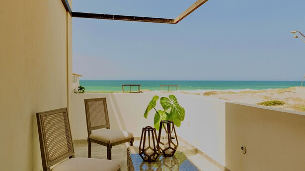 Un balcón cubierto con vista a la playa, dos sillones de mimbre, una mesita con una planta y piso de baldosas.