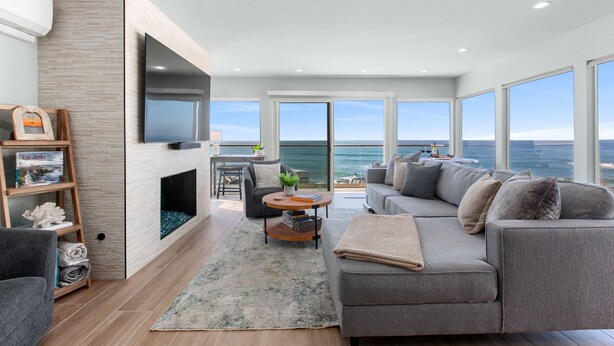A modern living room with a fireplace, a large flat-screen TV, a grey sofa, and a view of the ocean.