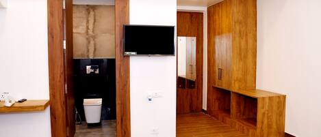 A modern hotel room with wooden flooring, a built-in bench, a flat-screen TV mounted on the wall, and a small kitchenette area.