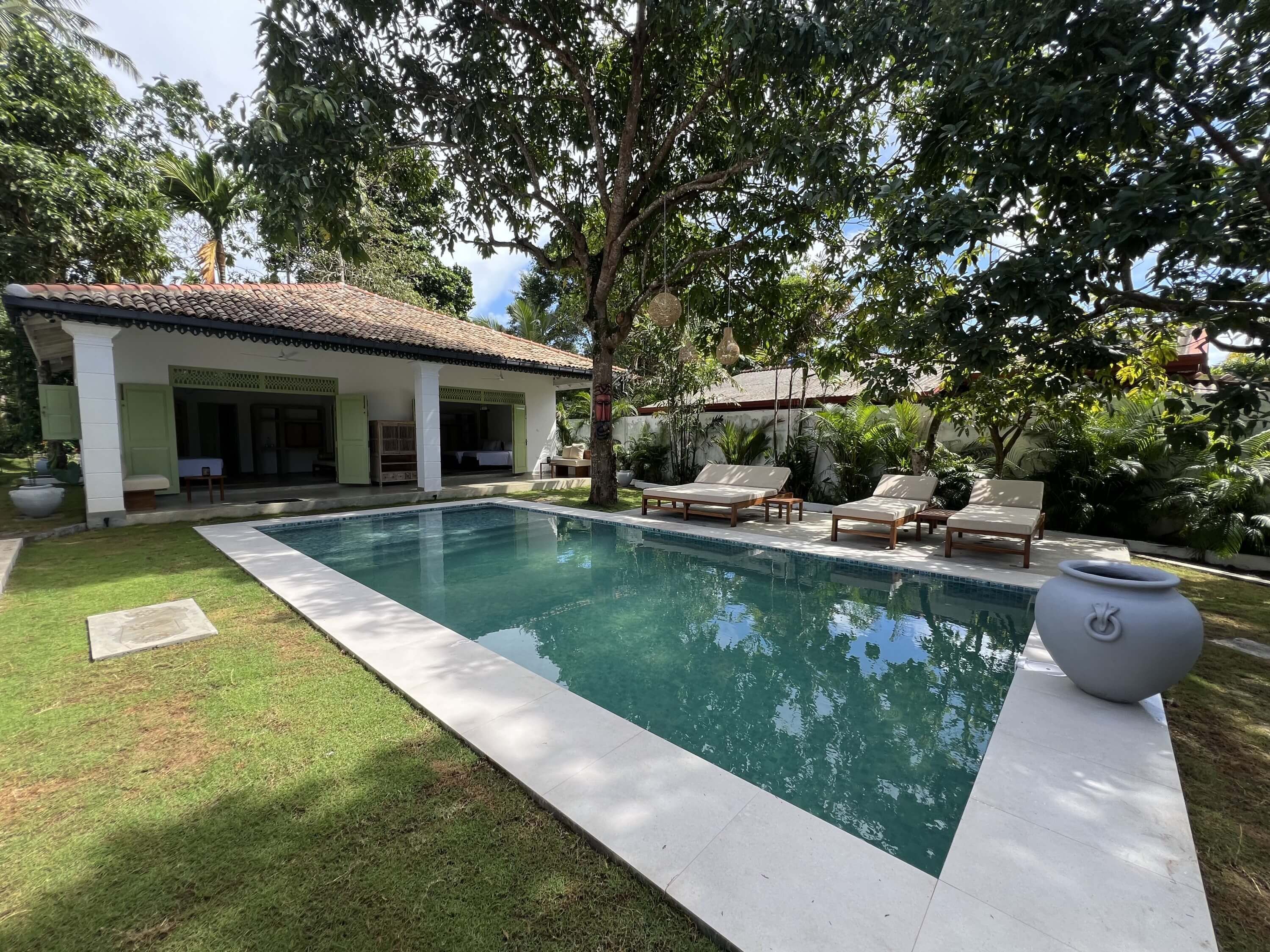 A swimming pool with lounge chairs, a covered patio, and lush greenery.