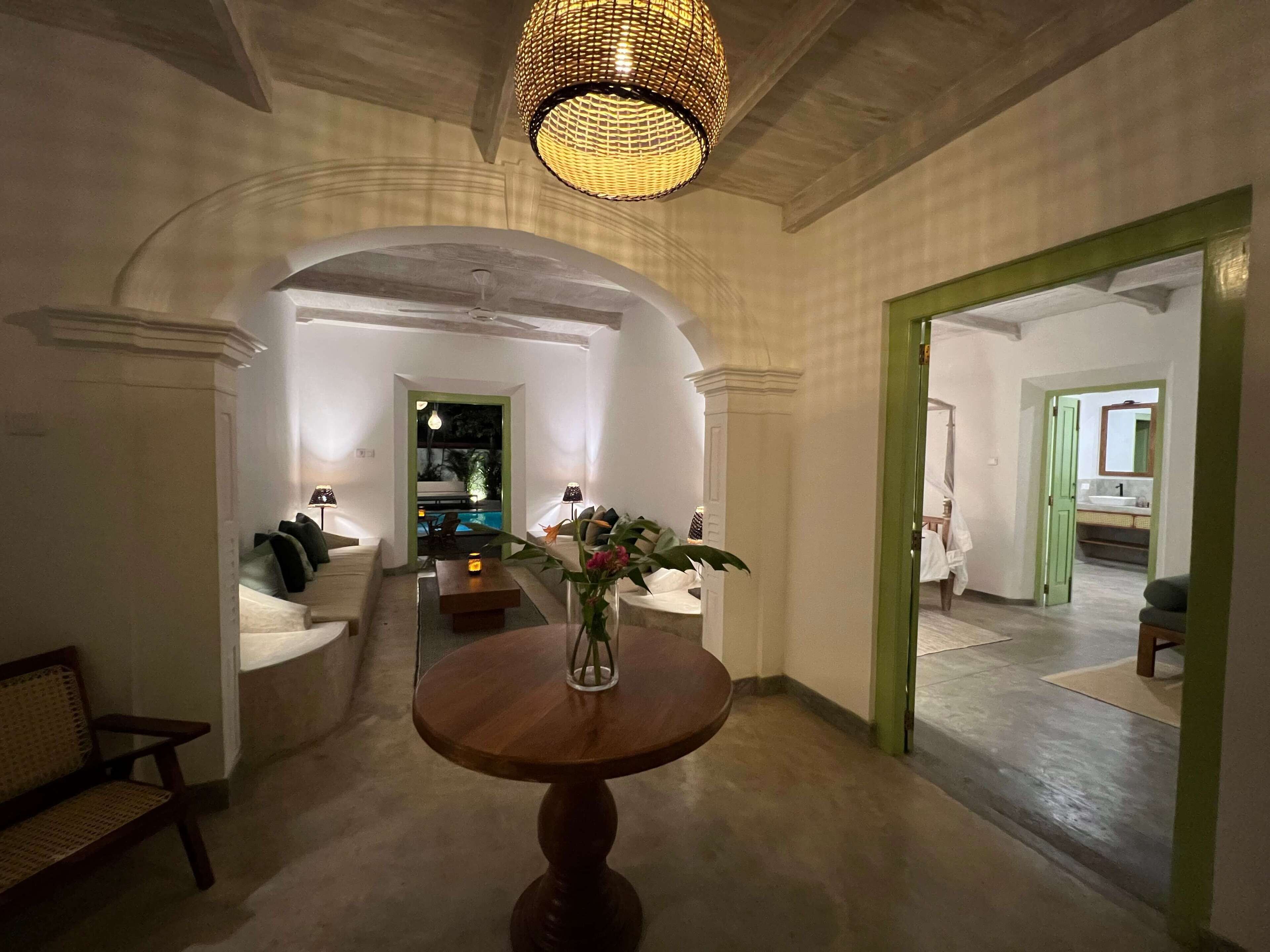 A hallway with a round wooden table, a wicker lamp, and a view into a dining area with a pool.
