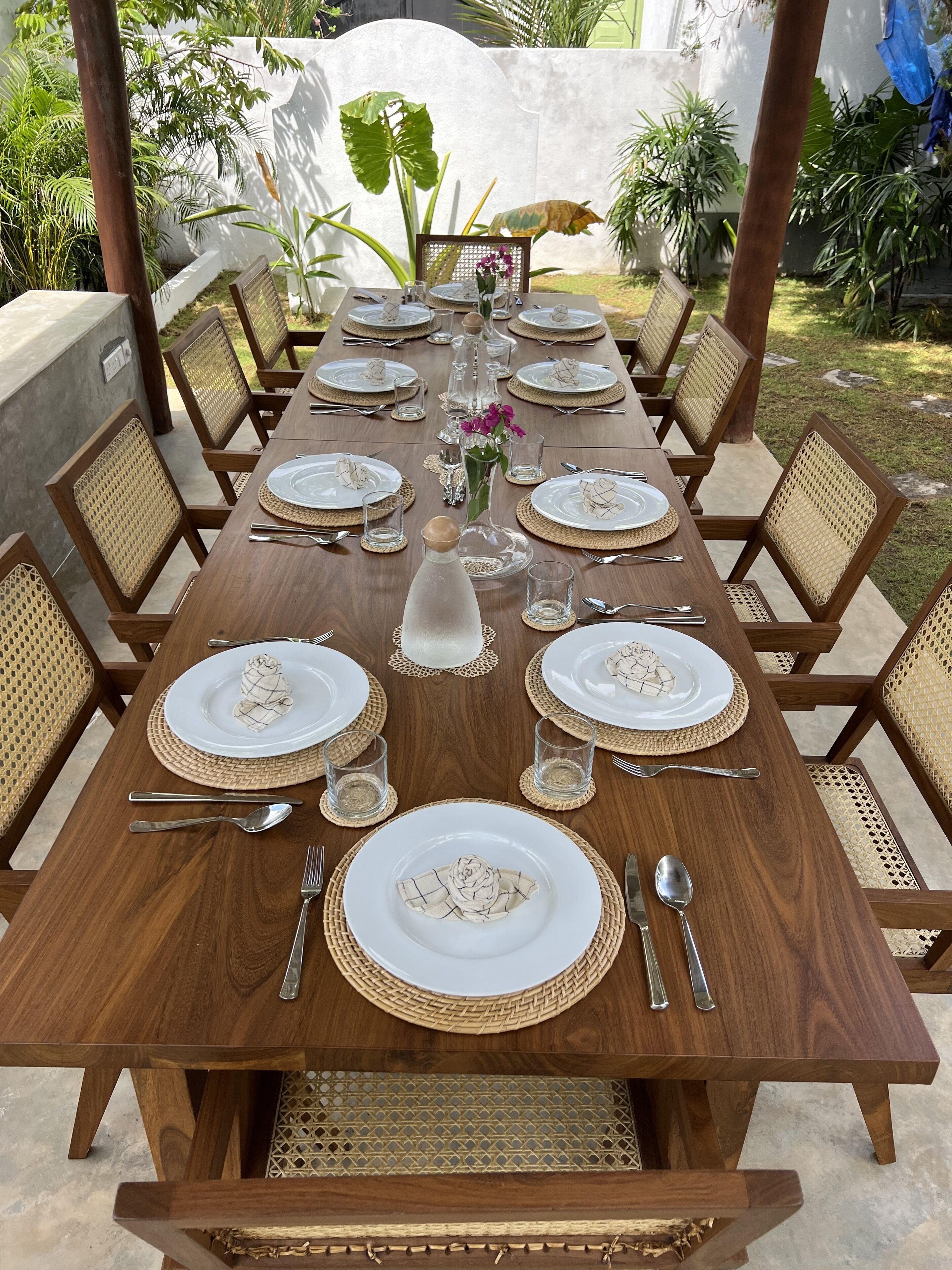 A wooden dining table set for eight with woven placemats and white plates with gold detailing.