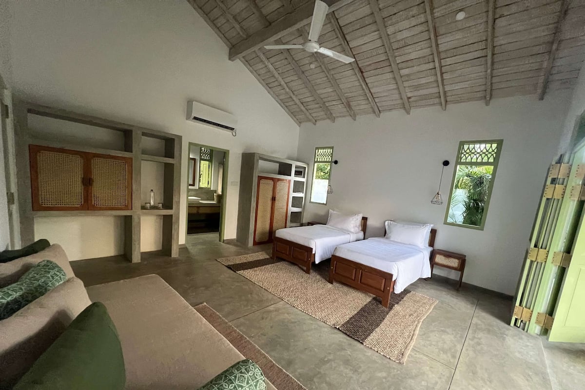 A room with two wooden beds, a sofa, a ceiling fan, and a bathroom visible through an open door.