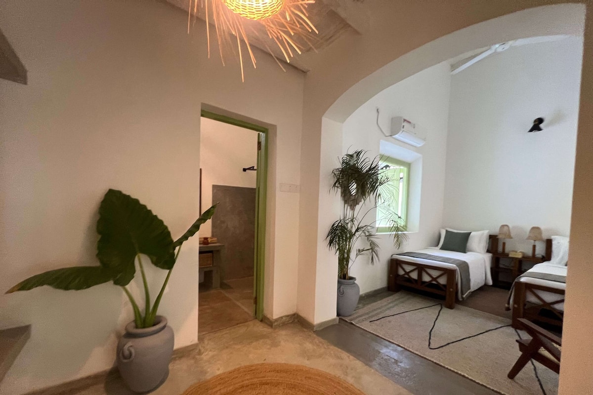 A cozy hotel room with a bed, a small table, a potted plant, and a unique ceiling light fixture.