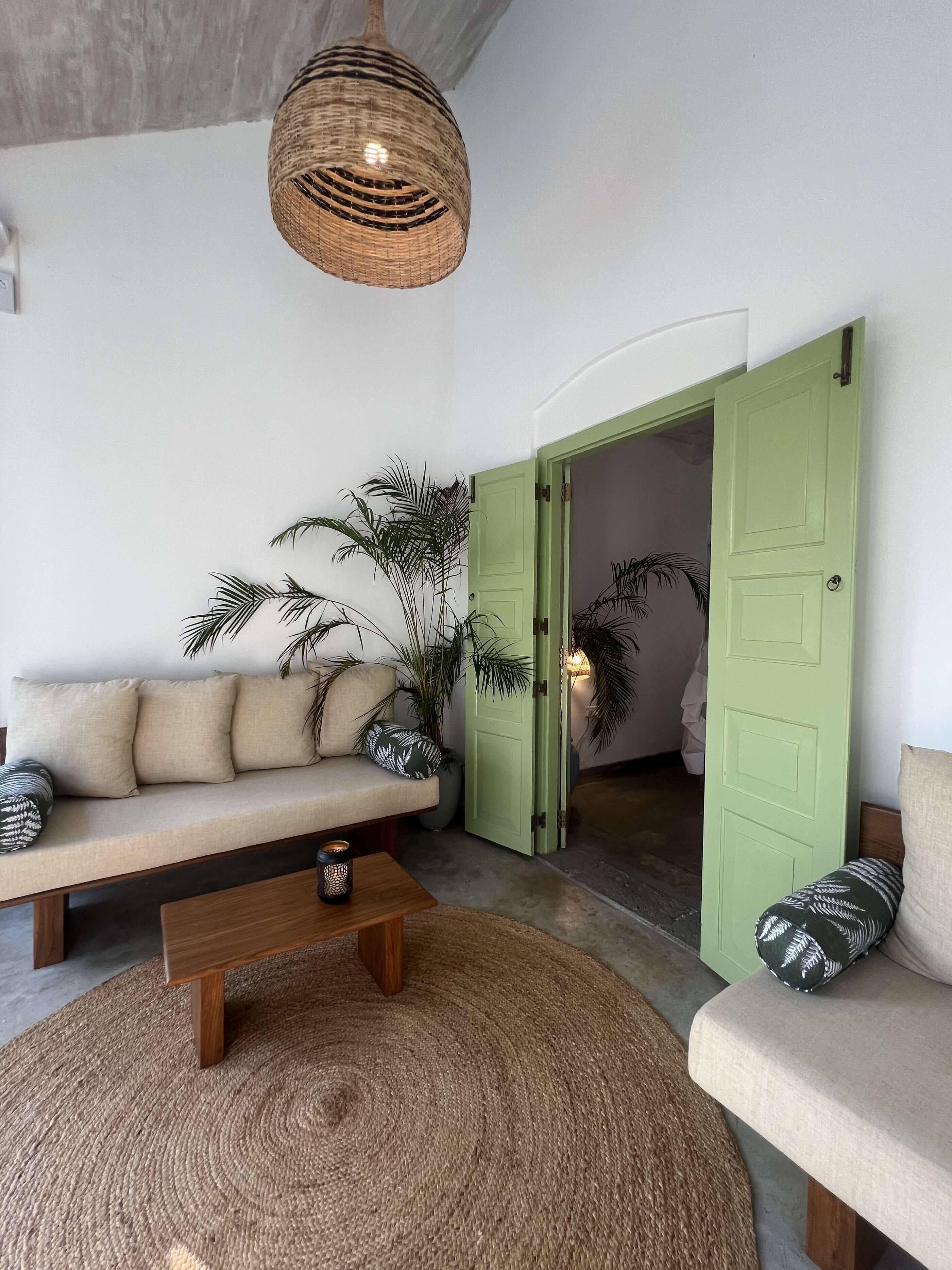 Een woonkamer met een bank, een salontafel, een hanglamp en een groene deur die naar een andere kamer leidt.