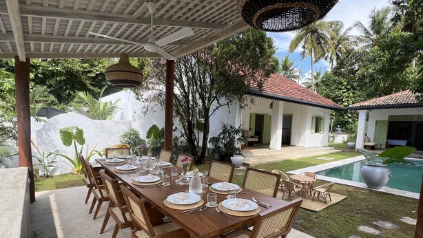 Un área de comedor al aire libre con una larga mesa de madera dispuesta para una comida, rodeada de sillas de mimbre, bajo un porche cubierto con ventilador de techo y lámparas colgantes.