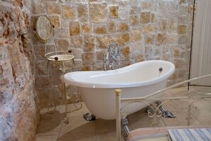 Une salle de bain avec une baignoire sur pieds, une petite commode avec un miroir et un mur en pierre.