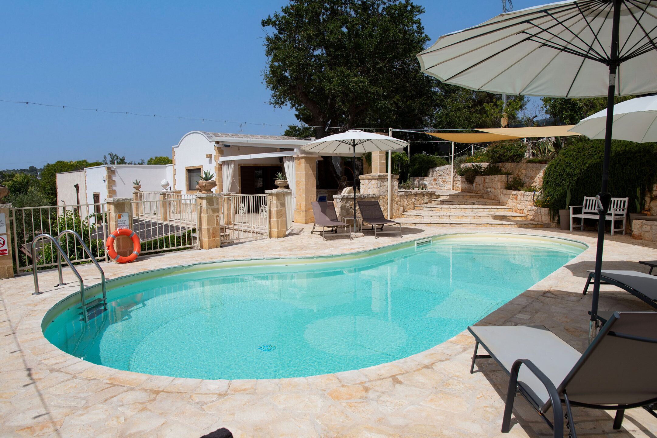 A swimming pool with lounge chairs and umbrellas, surrounded by stone walls and a building with a white facade.