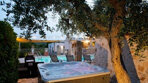A hot tub surrounded by outdoor seating and a pool, with stone walls and a tree in the background.