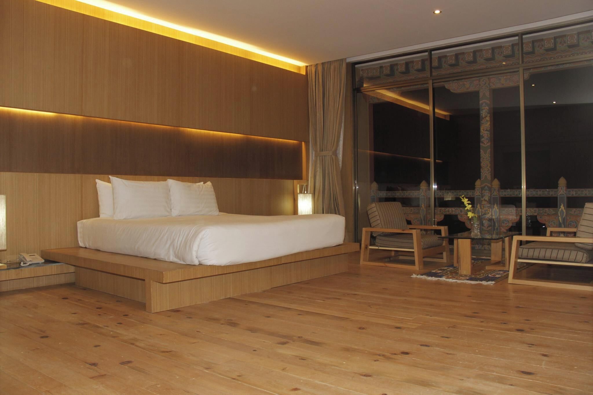 A modern hotel room with a wooden bed, a nightstand, a small table with a vase, two chairs, and a large window with a view of the night sky.