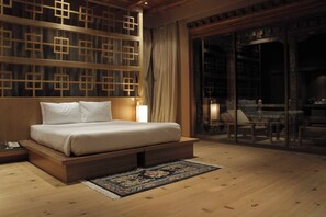 A bedroom with a wooden bed, a nightstand, a large window with curtains, and a view of a dining area. - Udumwara Resort and Spa (Paro)
