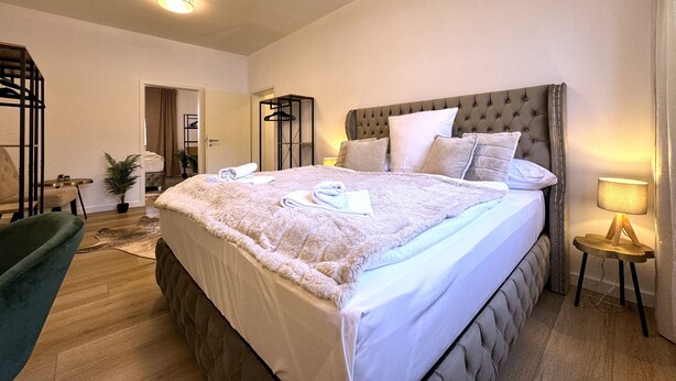 A spacious bedroom with a large bed, a chair, a small table, and a potted plant. - Dein Citynest - FeWo Meier (Bielefeld)