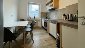 A modern kitchen with a dining area, wooden cabinets, and a window with a view of buildings. - Dein Citynest - FeWo Meier (Bielefeld)