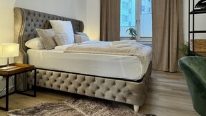 A bedroom with a tufted headboard, a bedside table with a lamp, a large bed with a white comforter, and a view of the street through a window with curtains. - Dein Citynest - FeWo Meier (Bielefeld)
