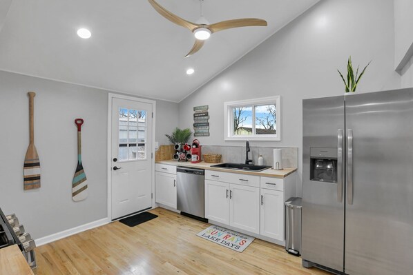 A modern kitchen with white cabinets, a stainless steel refrigerator, a ceiling fan, and a window with a view of the outdoors. - Homesmith Cottage|Hot Tub| Walk to Buckeye Lake (Buckeye Lake)