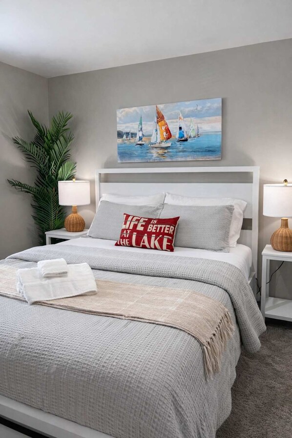 A neatly made bed with a pillow that reads "Life is better at the lake," a painting of sailboats on the wall, and a potted plant beside the bed.