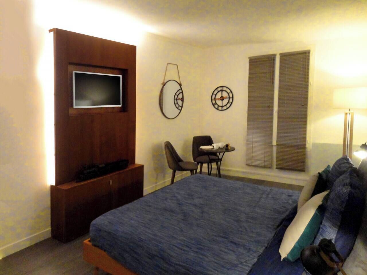 A hotel room with a bed, a TV mounted on a wooden panel, a desk with a chair, and a window with blinds.
