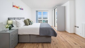A modern bedroom with a bed, bedside table, lamp, and a cityscape view through a large window. - Skyvillion 2 Bed Apt London Excel  O2 Arena (London)