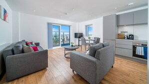 A modern living room with grey sofas, a dining table, and a kitchen area. - Skyvillion 2 Bed Apt London Excel  O2 Arena (London)