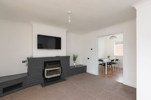 A living room with a fireplace, a flat-screen TV mounted above it, and a dining area with a table and chairs. - Skyvillion - 3bed Home W grdn For A Family Getaway (Stevenage)