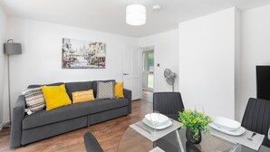 Una sala de estar moderna con un sofá gris, una mesa de comedor con sillas, un ventilador y una fotografía enmarcada de un paisaje urbano en la pared.