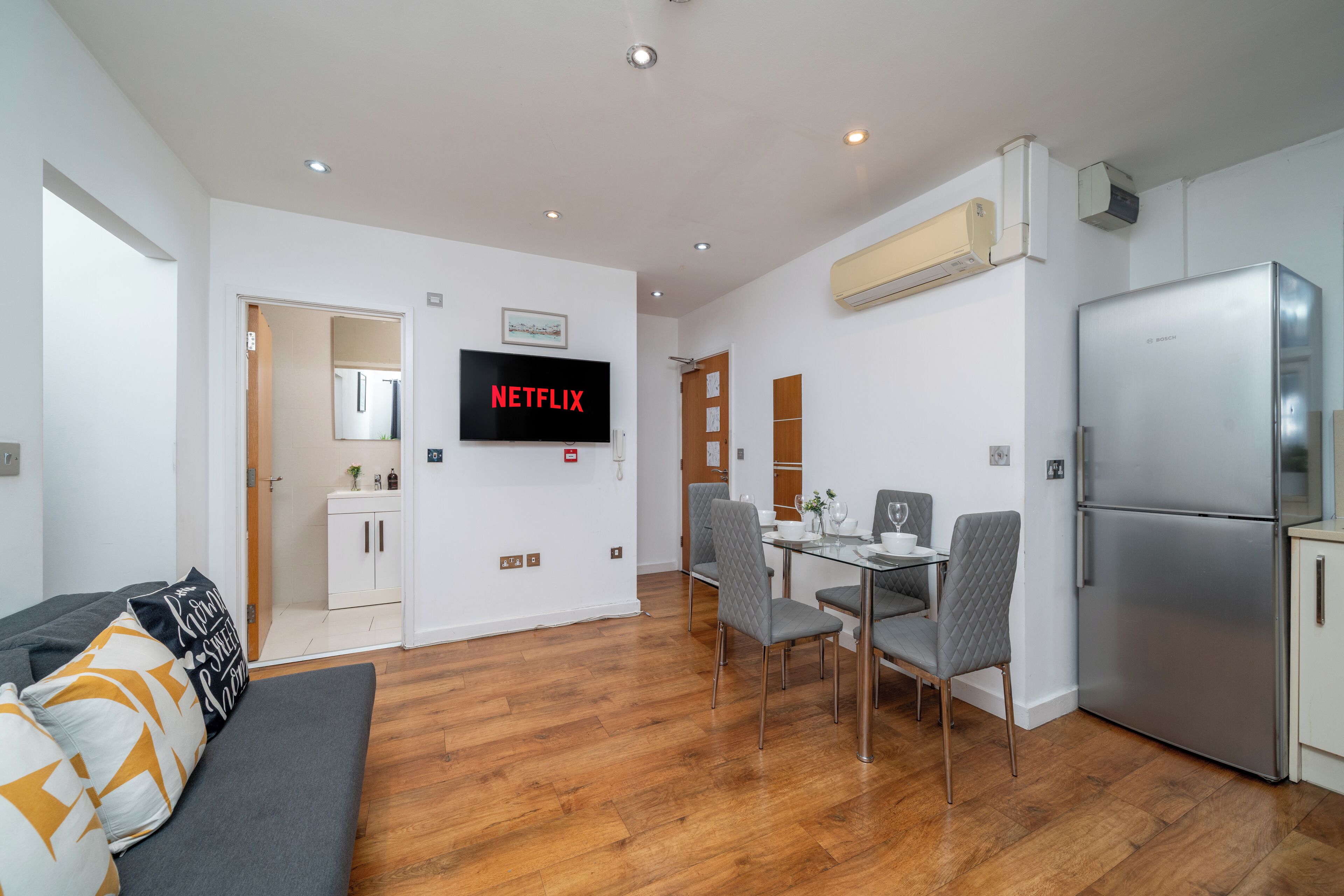 A modern living room with a flat-screen TV mounted on the wall, a dining area with a glass table and chairs, and a kitchen with a stainless steel refrigerator.
