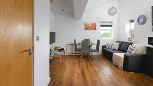 A modern living room with a black sofa, a glass coffee table, and a dining area with a table and chairs. - Skyvillion - Central Ldn 3bed Flat Ladbroke Grove (London)