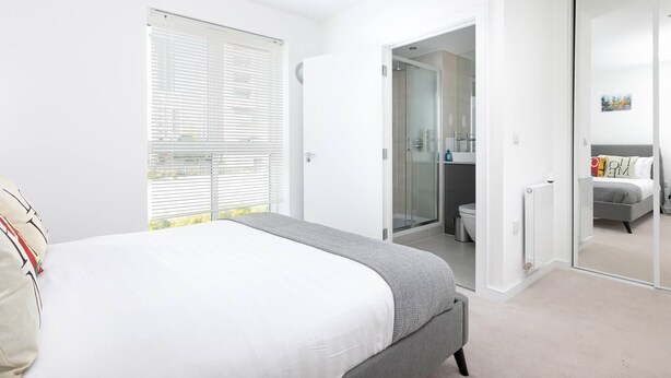 A bedroom with a bed, a window with blinds, a bathroom with a shower, and a closet with a mirror. - Skyvillion - Olympic Flat-excel o2 Arena westfield (London)