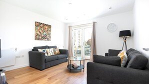A modern living room with two grey sofas, a glass coffee table, a black floor lamp, and a city view through a glass door. - Skyvillion - Olympic Flat-excel o2 Arena westfield (London)