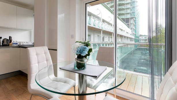 A modern kitchen with white cabinetry, a glass dining table with chairs, and a view of a building with balconies through a large window.