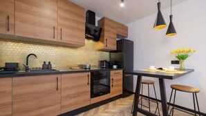 Una cocina moderna con armarios de madera, encimera negra, horno integrado y refrigerador negro. Hay dos lámparas colgantes negras y un florero con flores amarillas en la isla.