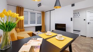 Una habitación de hotel moderna con una mesa de comedor, individuales amarillos, un jarrón con flores amarillas, un sofá, un televisor y una puerta blanca.