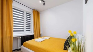 Un dormitorio con cama, ventana con persianas, radiador y un jarrón con flores amarillas.